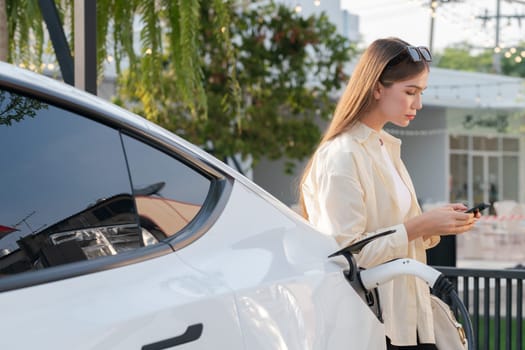 Sustainable urban commute with young woman pay electricity for EV electric car recharging at outdoor cafe in springtime garden, green city sustainability and environmental friendly EV car. Expedient