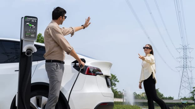 Young couple recharge EV car battery at charging station connected to power grid tower electrical industrial facility as electrical industry for eco friendly vehicle utilization. Expedient