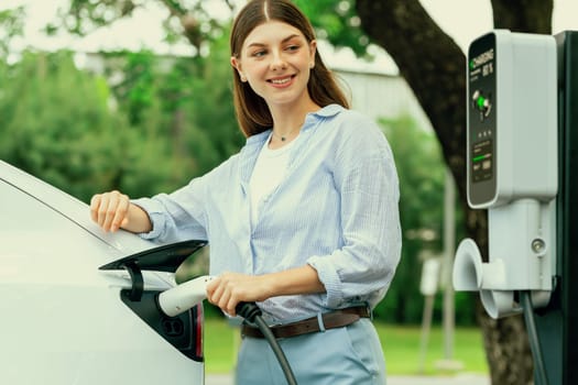 Young woman recharge EV electric vehicle's battery from EV charging station in outdoor green city park scenic. Eco friendly urban transport and commute with eco friendly EV car travel. Exalt