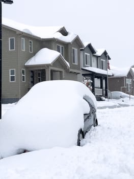 A car is buried under a thick layer of snow in front of a row of townhouses also draped with snow, presenting a challenging winter scene.