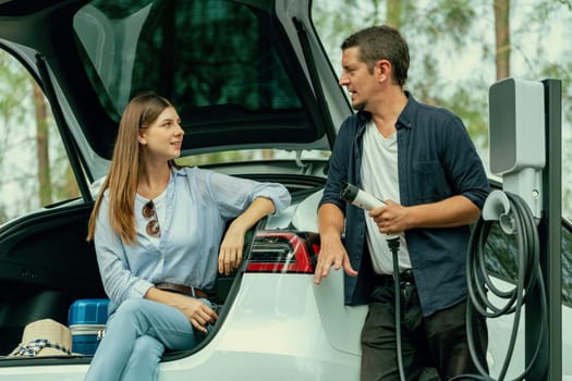 Lovely young couple recharging battery for electric car while relaxing and sitting on the trunk during road trip travel EV car in autumnal forest. Eco friendly travel on vacation during autumn. Exalt