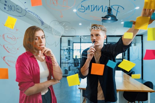 Couple of businesspeople discussing and brainstorming about solving marketing problem in front of glass board at business meeting by using mind map and sticky notes. Working together. Immaculate.