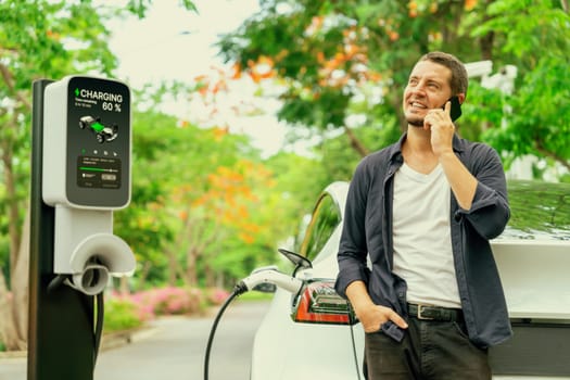 Man talking on smartphone while recharging electric car battery charging from EV charging station during vacation holiday road trip at national park or autumnal forest. Exalt