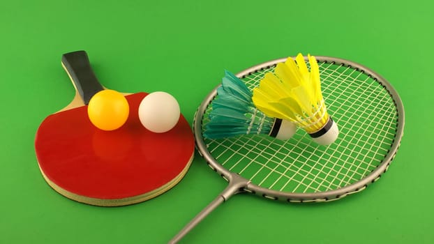 Table tennis racket with a yellow ball and a white ball on it and badminton racket with a feather shuttlecock on clear green background