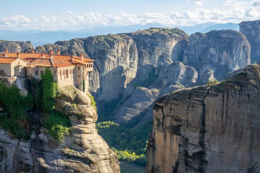 Greece. Sunny summer day in Kalambaka. Rock monastery with red roofs. Light clouds in the blue sky over the mountain range in the background