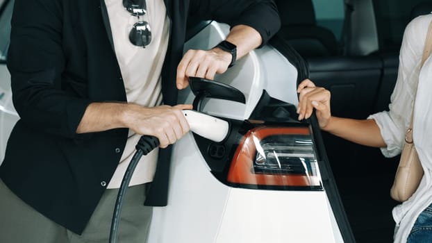 Young couple travel with EV electric car to shopping center parking lot charging in downtown city showing urban sustainability lifestyle by green clean rechargeable energy of electric vehicle innards