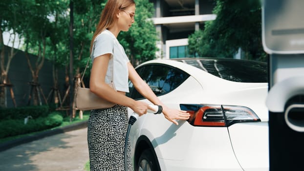 Young woman recharge EV electric car battery at residential area EV charging station in urban city condo with sustainable green and renewable clean energy lifestyle for electric vehicle innards
