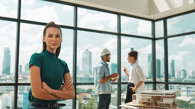 Young beautiful interior designer crossing arms while engineer team talking about house design. Skilled businesswoman smiling at camera with confident while sitting near house model. Tracery