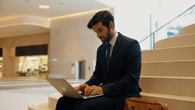 Skilled investor working or planing strategy by using laptop at stair. Professional business man wearing suit while working and typing data analysis by using laptop at modern hotel. Exultant.