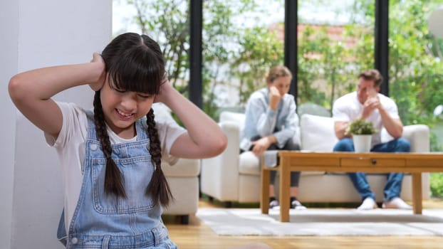 Stressed and unhappy young girl huddle in corner, cover her ears blocking sound of her parent arguing in background. Domestic violence at home and traumatic childhood develop to depression. Synchronos