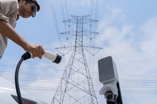 Man recharge EV electric car battery at charging station connected to electrical power grid tower on sky background as electrical industry for eco friendly vehicle utilization. Expedient