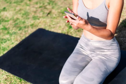 unrecognizable young woman in sportswear kneeling on her mat using mobile phone for a yoga session on the park grass, technology and healthy lifestyle concept