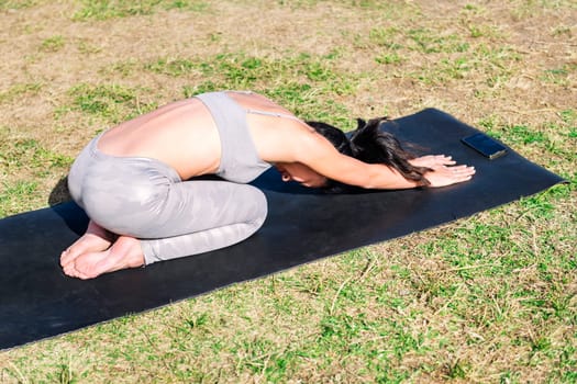 young woman in sportswear kneeling on her yoga mat doing back stretching exercises on the grass in the park, active and healthy lifestyle concept