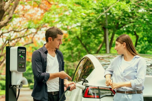 Lovely young couple recharging battery for electric car during autumnal road trip travel EV car in autumnal forest. Eco friendly travel on vacation during autumn. Exalt