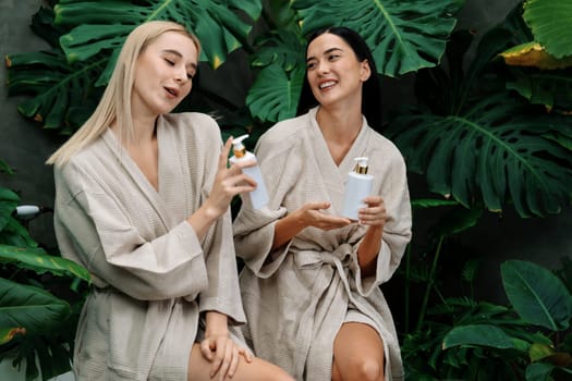 Tropical and exotic spa garden with bathtub in modern hotel or resort with two women in bathrobe holding beauty skincare product while enjoying leisure lush with greenery foliage background. Blithe