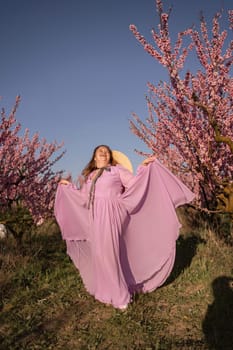 Woman blooming peach orchard. Against the backdrop of a picturesque peach orchard, a woman in a long pink dress and hat enjoys a peaceful walk in the park, surrounded by the beauty of nature