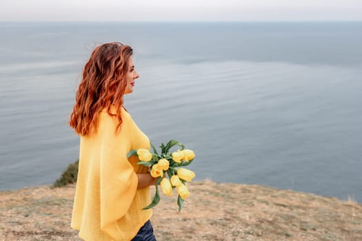 Rear view of a woman with long hair against a background of mountains and sea. Holding a bouquet of yellow tulips in her hands, wearing a yellow sweater.