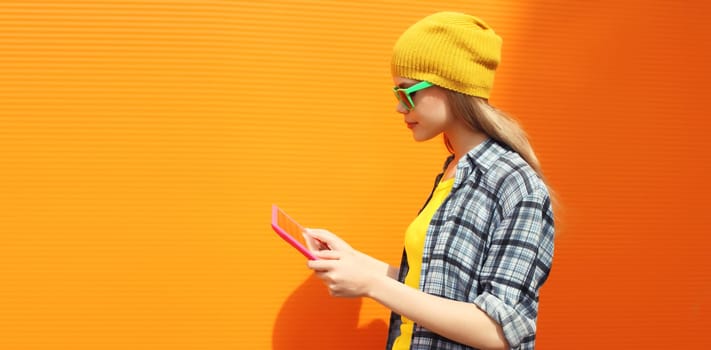 Modern stylish young woman using digital tablet computer looking at device in colorful clothes on city street on orange wall background