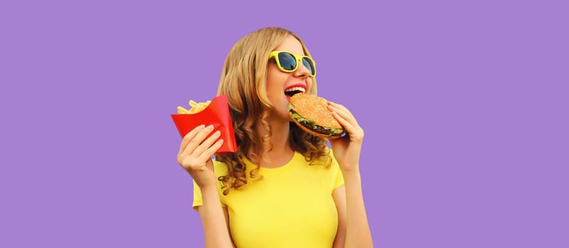 Portrait of happy cheerful young woman eating burger fast food and french fries, fried potatoes isolated on purple studio background
