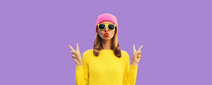 Portrait of stylish modern young woman blowing kiss posing in colorful clothes, yellow sweater, pink hat on purple studio background