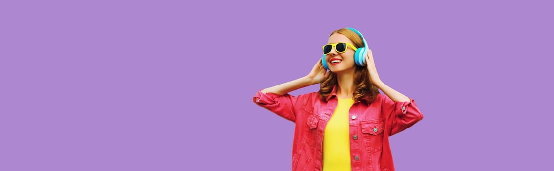 stylish modern happy young woman listening to music with headphones on colorful purple studio background, blank copy space for advertising text