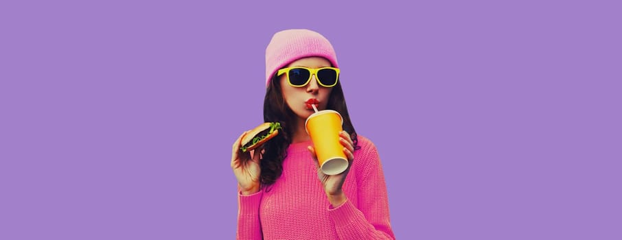 Portrait of stylish modern young woman drinking juice eating burger fast food on colorful purple studio background
