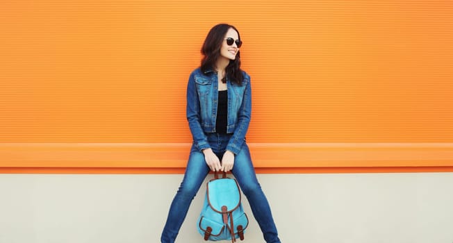 Stylish happy smiling brunette young woman with backpack posing on city street on orange wall background