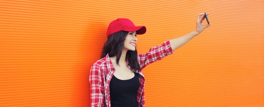 Summer portrait of happy smiling young woman taking selfie with mobile phone in red baseball cap on colorful orange background on city street