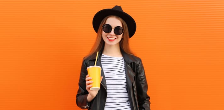 Portrait of stylish young woman drinking fresh juice or coffee holding cup in black rock style leather jacket, round hat on colorful orange background