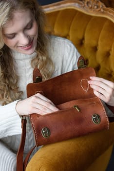 beautiful curly blond hair woman posing with a small tube brown bag in a vintage chair. Model wearing stylish white sweater and classic trousers