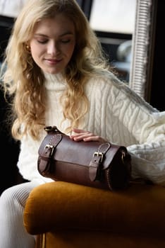 beautiful curly blond hair woman posing with a small tube brown bag in a vintage chair. Model wearing stylish white sweater and classic trousers