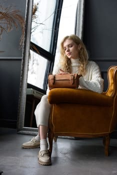 beautiful curly blond hair woman posing with a small tube brown bag in a vintage chair. Model wearing stylish white sweater and classic trousers