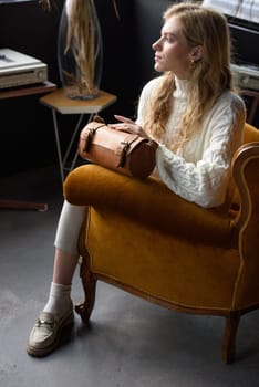 beautiful curly blond hair woman posing with a small tube brown bag in a vintage chair. Model wearing stylish white sweater and classic trousers