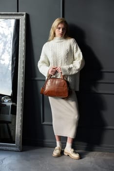 Studio portrait of beautiful woman with a curly blond hair holding brown bag, posing on gray background. Model wearing stylish sweater and skirt