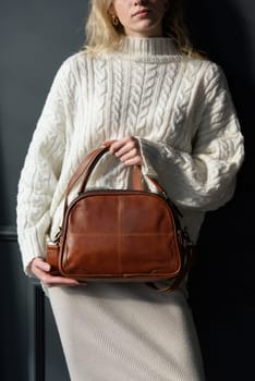 Studio portrait of beautiful woman with a curly blond hair holding brown bag, posing on gray background. Model wearing stylish sweater and skirt