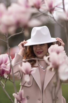 Woman magnolia flowers, surrounded by blossoming trees, hair down, white hat, wearing a light coat. Captured during spring, showcasing natural beauty and seasonal change