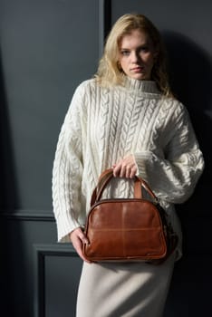 Studio portrait of beautiful woman with a curly blond hair holding brown bag, posing on gray background. Model wearing stylish sweater and skirt