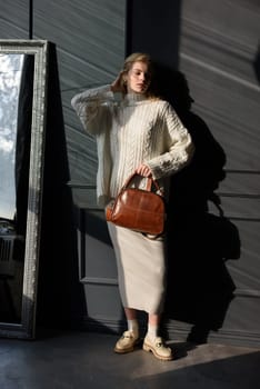 Studio portrait of beautiful woman with a curly blond hair holding brown bag, posing on gray background. Model wearing stylish sweater and skirt