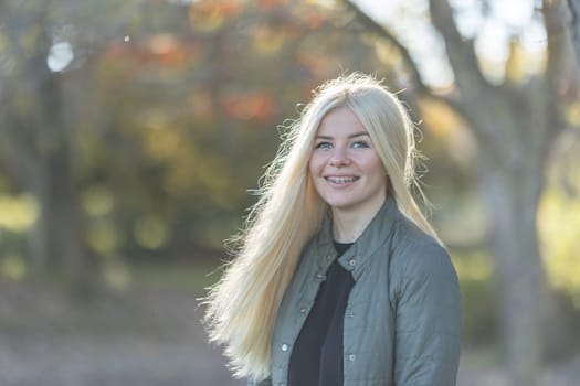 A young woman with long blonde hair is standing in front of trees, smiling. She has braces and seems to enjoy the park setting.