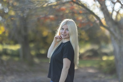 A young woman with braces smiles while standing in front of a cluster of trees in a park on a sunny day.