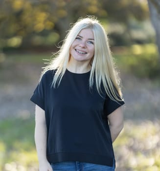 A young woman with blonde hair and braces, smiling brightly while wearing a black shirt in a park setting.