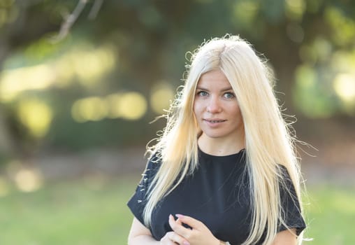 A young woman with braces, standing in a park with long blonde hair, smiling.