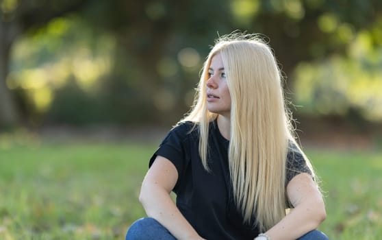 A young woman with long blonde hair and braces is sitting on the grass in a park, smiling.