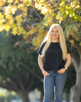 A young woman with braces stands confidently in a park, with her hands on her hips in front of a tree.