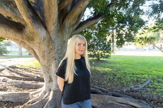 A young woman with braces is standing in front of a tree in a park, smiling.