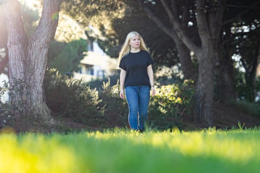 A young woman with braces is smiling as she walks through green grass in front of several tall trees in a park setting.