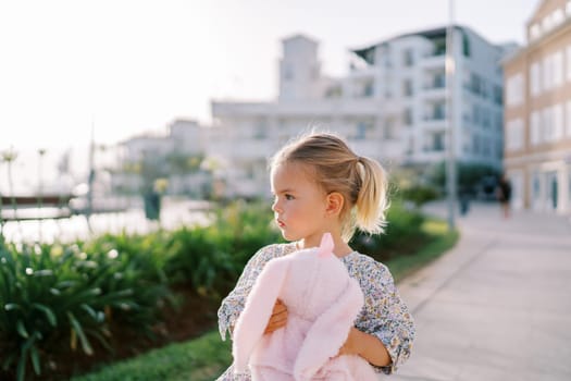 Little girl with a pink plush rabbit in her hands walks along the yard of a multi-story building and looks to the side. High quality photo