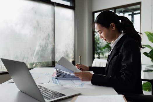 A woman is writing on a piece of paper with a pen. She is wearing a black suit and is sitting at a desk. The paper has a lot of numbers and calculations on it