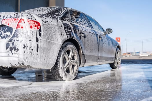 A vehicle is engulfed in soapy foam during a car wash, covering the tires, wheels, hood, and entire exterior of the car. Self-service car wash