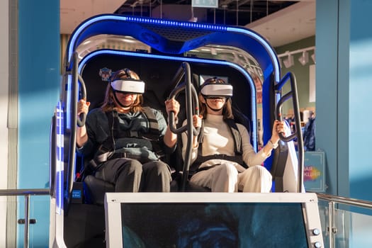 Moscow, Russia, March 14, 2024. Two woman is sitting in an electric blue virtual reality chair, wearing a headset. The machine combines engineering and fun in a display device